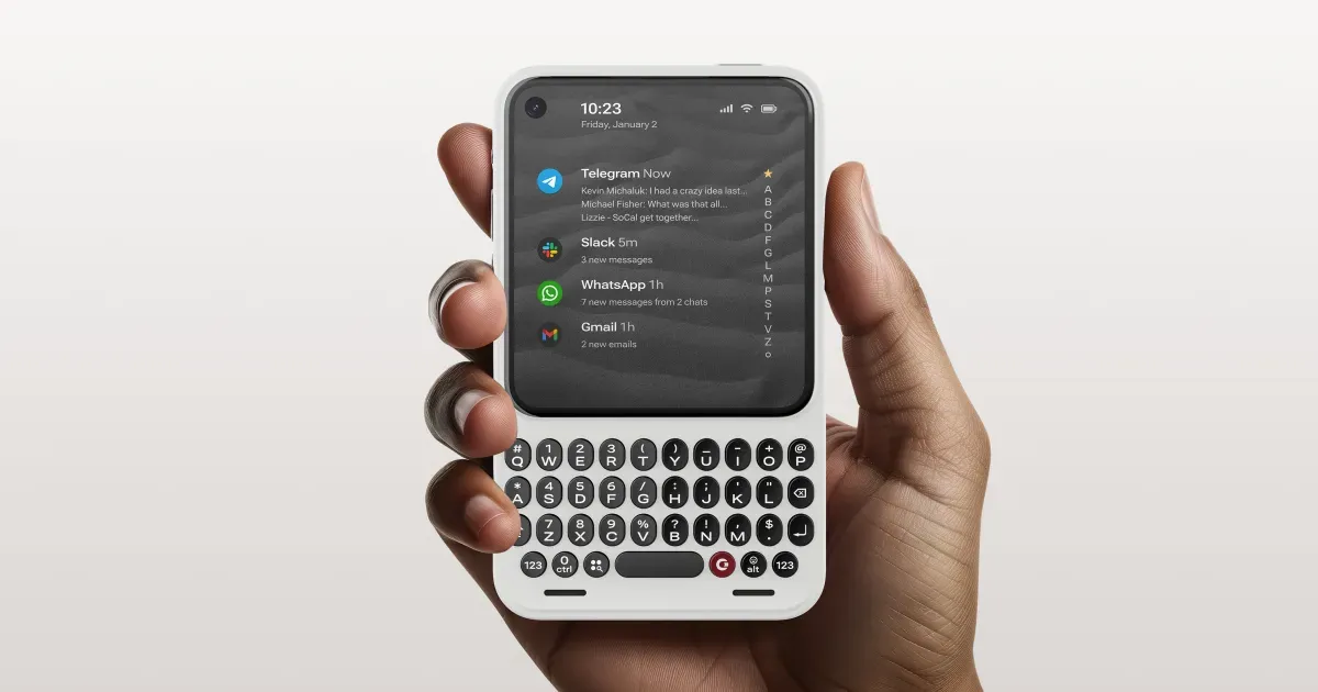 A hand holding the white Clicks Communicator, its screen showing a list of messages above a rounded QWERTY keyboard