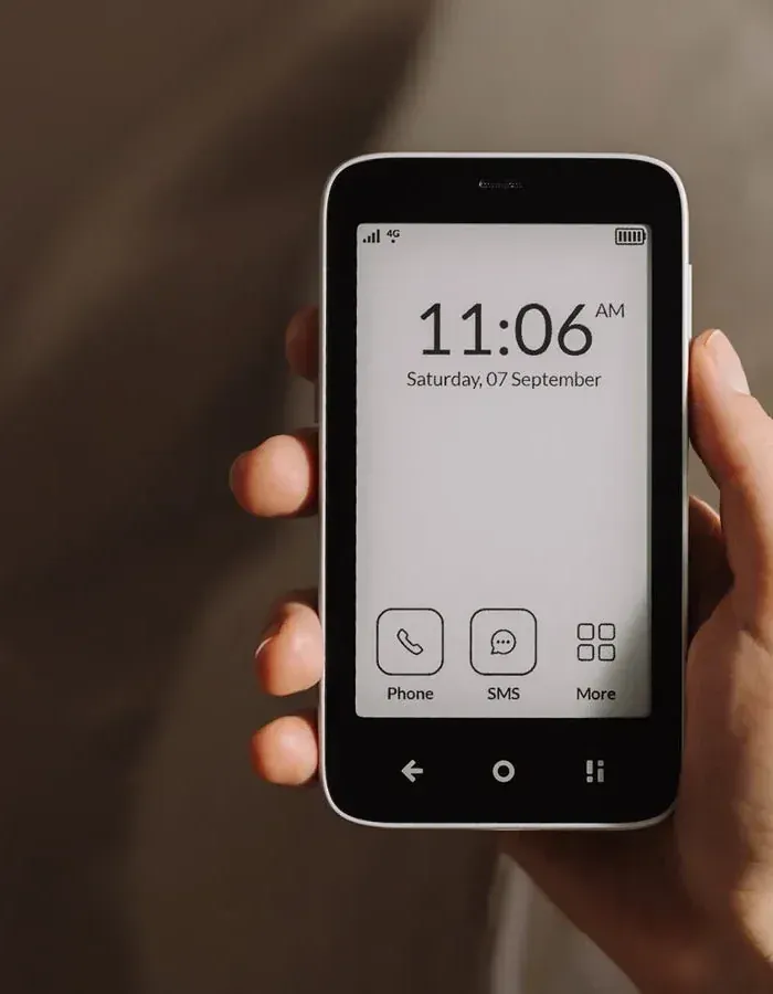 A hand holding the Mudita Kompakt, its E Ink screen showing the time and three buttons: Phone, SMS, More